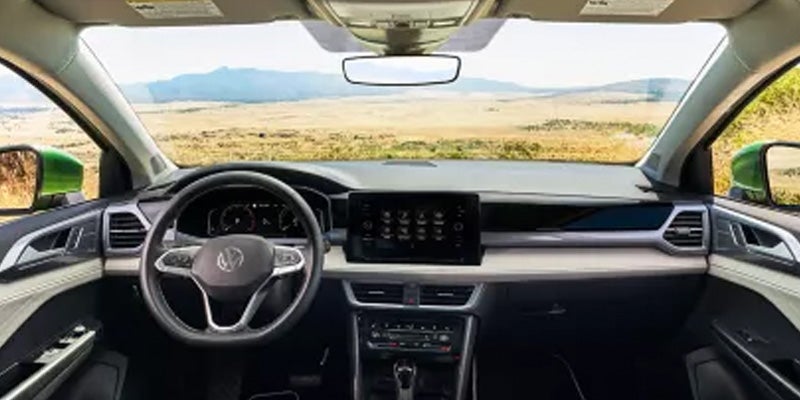 The interior of a Volkswagen car dashboard with a steering wheel, screen, and tan landscape visible through the windshield.